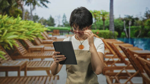 Woman in apron adjusts glasses while holding a tablet on a poolside deck among wooden lounge chairs; concentration hospitality.