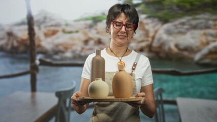 Woman in apron holding ceramic vases on a tray with hands visible, grimacing while presenting pottery in a studio; unease craft.