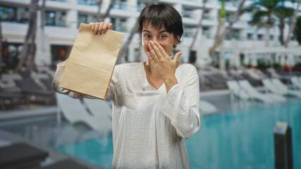 Woman holding a paper bag with one hand raised and the other hand on her chest, smiling by a hotel...