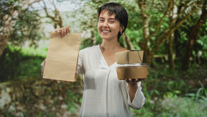 Woman with short hair holding a paper bag and stacked cardboard food boxes, smiling and holding...