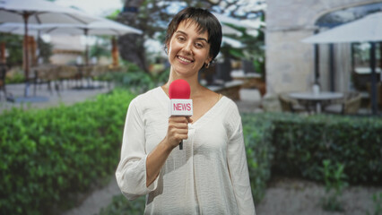 Fototapeta premium Woman holding red news microphone and smiling while reporting at a building courtyard with umbrellas, outdoor tables and hedges, wearing white blouse and short hair; confidence.