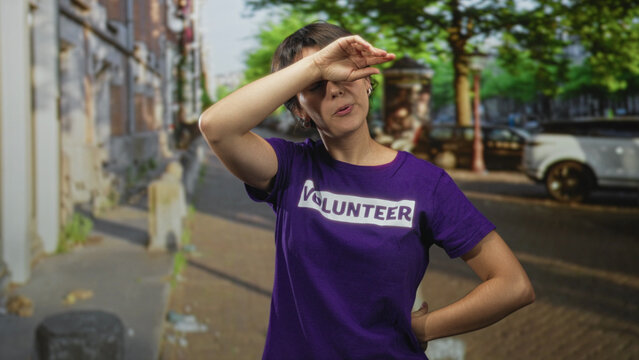 Woman in volunteer tshirt standing with hands on hips and wiping forehead on a tree lined street near parked car and sidewalk; determination.