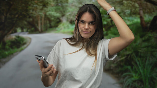 Young hispanic woman holding smartphone in outdoor park setting looks surprised with green trees around her