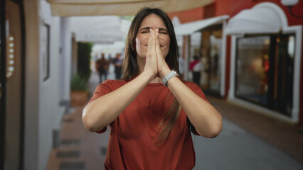 Young hispanic woman in red shirt prays with eyes closed on a bustling city street filled with shops and people, exuding hope and peace in a vibrant urban setting.