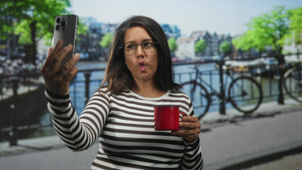 Brunette woman taking selfie with smartphone while holding bright red mug on urban street canal walkway with railing and bicycle in view; playful.