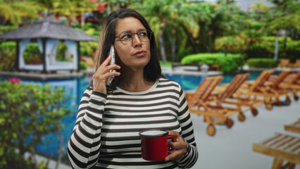 Woman holds phone to ear and a red mug, wearing a striped top by a resort pool with wooden lounge chairs and a gazebo; serenity and focus.