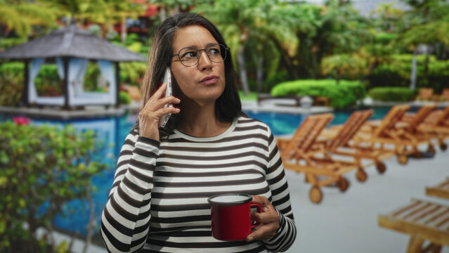 Woman holds phone to ear and a red mug, wearing a striped top by a resort pool with wooden lounge chairs and a gazebo; serenity and focus.