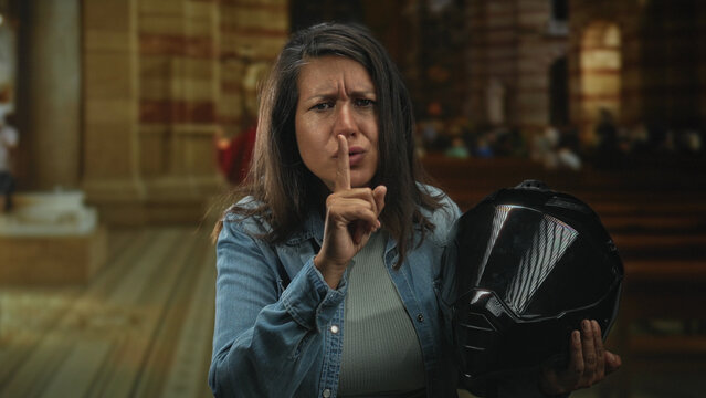 Middle age woman holding a glossy black motorcycle helmet, finger to lips for silence while standing inside a church nave with wooden pews and stone columns; quiet contemplation.