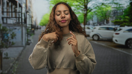 Woman touching curly hair and twirling a lock with both hands on a city street, eyes closed and adjusting curls; quiet contemplation.