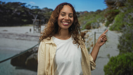 Young african american woman smiling, finger pointing to right on sandy seaside path with lifeguard tower visible; joy travel.