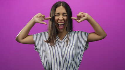 Hispanic young brunette woman covering her ears with fingers in a pink studio, striped shirt visible; frustration coping.