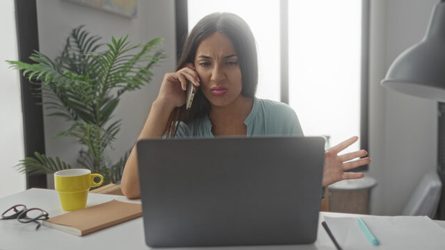 Hispanic woman holding smartphone to ear and typing on laptop at a desk in a bright home interior; work stress frustration.