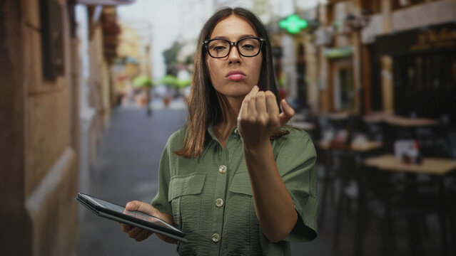 Woman holding tablet beckons with fingers toward viewer at street cafe terrace outdoors wearing glasses; impatience. - Powered by Adobe