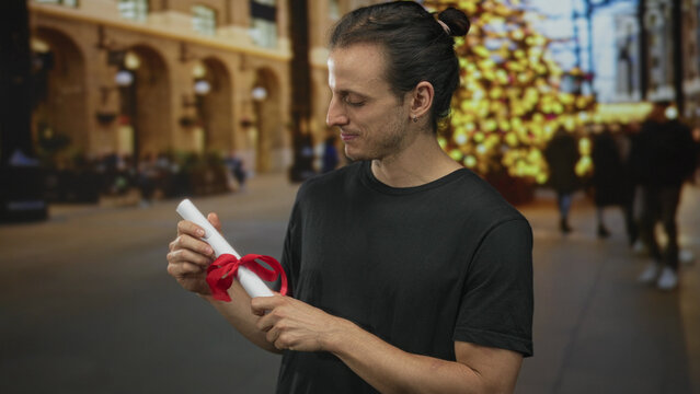 Man holds rolled certificate with red ribbon using both hands on street near decorated tree; quiet pride celebration.