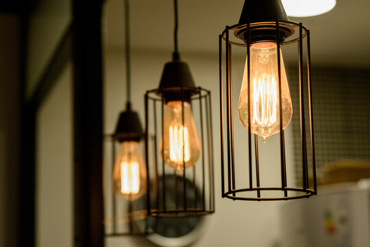 Fila de l&aacute;mparas colgantes estilo industrial con bombillas de filamento Edison encendidas. Iluminaci&oacute;n c&aacute;lida retro en jaulas de metal negro con enfoque selectivo y fondo oscuro.