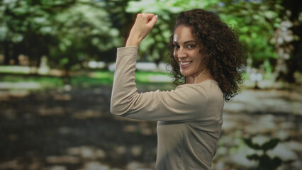 Woman with curly hair smiling and reaching her arm forward in a forest path, turning toward camera...