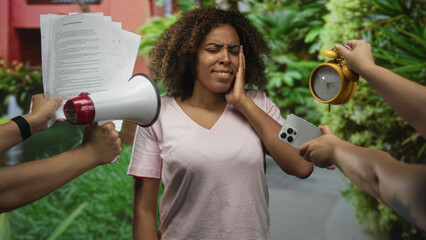 Young woman with a megaphone, papers, phone and alarm clock held near her while covering ears with hands on a street; stress.