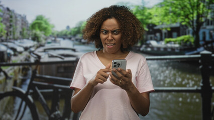 Woman holding smartphone points finger to phone on street in amsterdam canal scene with puzzled frown; digital frustration.