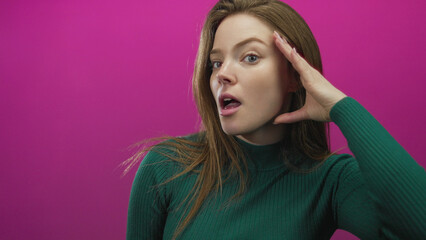 Woman in green sweater cups her hand to ear and opens mouth in pink studio with curious tilt of...