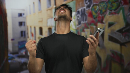 Young hispanic man outdoors using smartphone against graffiti wall expressing excitement and concentration with gestures
