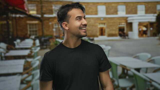 Young hispanic man smiling on a terrace with outdoor seating in a restaurant setting, enjoying a relaxed atmosphere under a sunny sky.