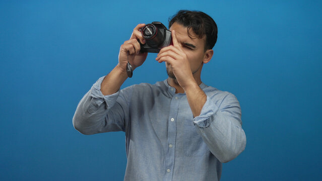 Young hispanic man thinking with camera against isolated blue background, wearing casual shirt, conveys creativity and contemplation in a studio setting.