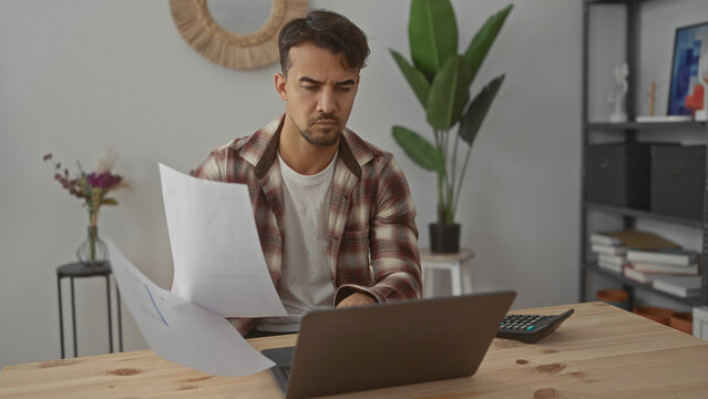 Young hispanic man working in an office holding papers with a focused expression beside a laptop and calculator, surrounded by indoor decor elements.