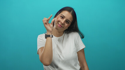 Woman pinching fingers by face wearing white t shirt and smartwatch in studio with teal backdrop;...