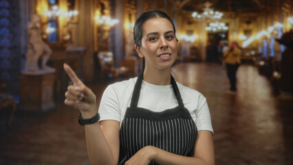 Young brunette woman wearing a striped apron points finger to object while standing in ornate museum building lobby; welcoming hospitality.