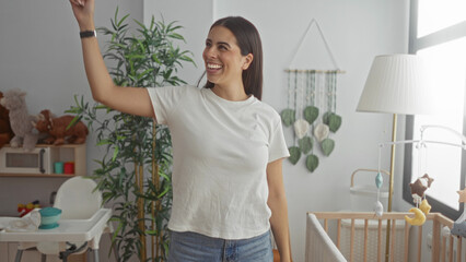 Woman laughing with raised arm by crib in bedroom, nursery mobile and toys visible near window; joy motherhood.