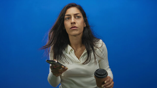 Young hispanic woman holding smartphone and takeaway coffee, squinting while checking phone in a blue studio; frustration.