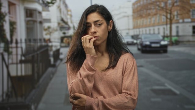 Woman biting nails with hand to mouth and arm folded on a street lined with townhouses and parked cars; worry.