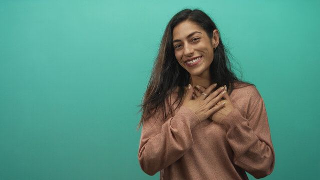 Woman smiling with hands pressed to chest wearing rust sweater in teal studio; gratitude warmth appreciation.