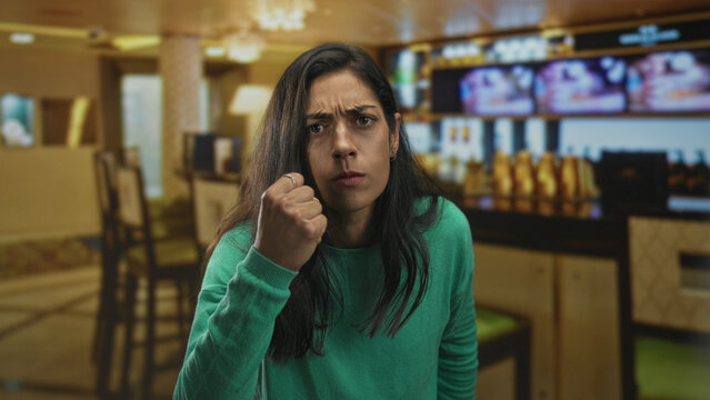 Woman clenches fist and glares in a restaurant building with raised hand near face and furrowed brow, leaning forward in an aggressive stance; anger confrontation.