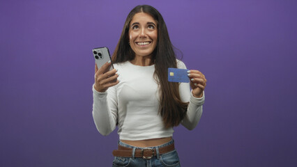 Young woman holds creditcard in one hand and smartphone in the other, hand visible, smiling in a studio; excitement.
