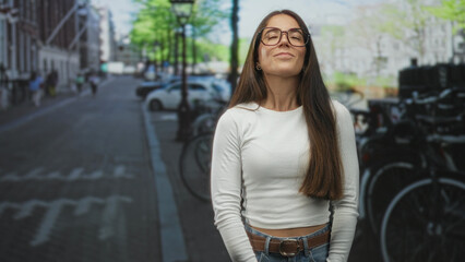 Woman wearing glasses smiles with eyes closed and hands loosely clasped at waist on a street beside parked bikes and cars; quiet contentment.