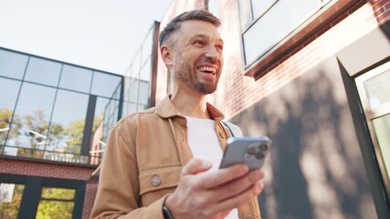 Caucasian male holding smartphone and smiling while swiping on touch screen. Man carrying backpack standing near reflective building wall. Engaged male browsing posts during relaxed moment.