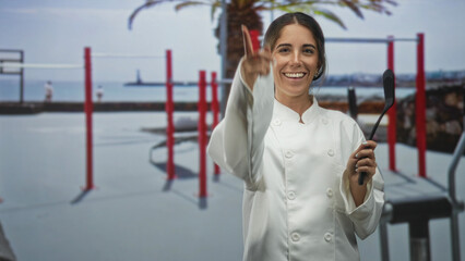 Chef woman in white coat holds spatula and gestures with open hand toward camera on a street by the...