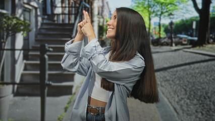 Young hispanic woman pointing index fingers upward while smiling on a cobblestone street with visible midriff and belt; joyful curiosity.