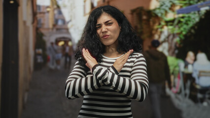 Fototapeta premium Woman with crossed forearms making an x gesture on a street amid cafe tables and pedestrians; rejection boundary assertiveness.