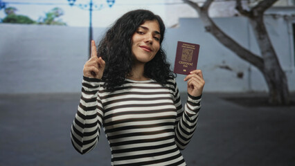 Woman points finger while holding czech passport on a city street, wearing striped top and smiling face; travel pride.