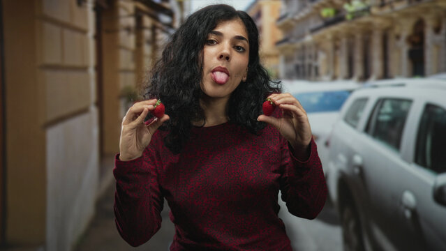 Young arab woman holding strawberries and sticking tongue out on a busy city street; playful summer freshness.