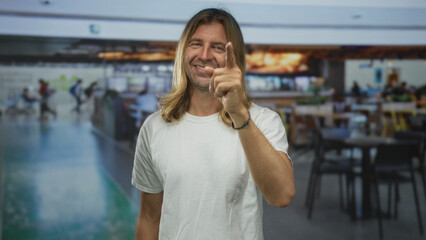 Man with long blond hair smiling and pointing finger at camera inside a busy airport terminal near...