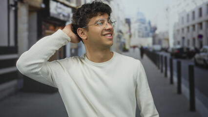 Man adjusts hair in street as he smiles wearing glasses before blurred urban building facades at...