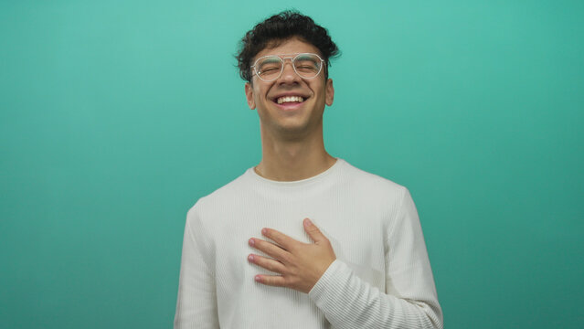 Young hispanic man wearing glasses with hand on chest laughing in studio setting; amusement gratitude.
