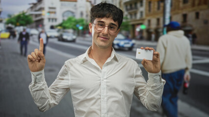 Man holding white chip card between thumb and forefinger on city street, wearing glasses and white shirt, presenting card; confidence.