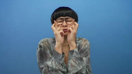 Woman crossing fingers with hopeful expression standing against a solid blue background wearing...