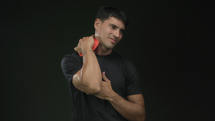 Young hispanic man pressing ice pack to neck in studio with dark green background; pain discomfort healing strain.