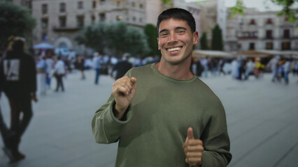 Man in olive green sweater dances with raised hands and smiling face on crowded city street; happiness.