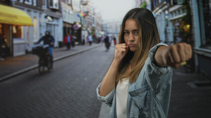 Young hispanic woman punching with fist toward camera in city street, defensive stance and focused expression; self defense defiance.